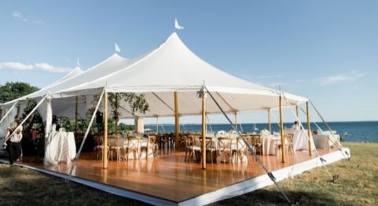 Outdoor event tent with open sides and banquet tables and chairs set up on a polished wood platform floor on the grounds of the UConn Avery Point campus. Views of the sea are visible in the background.