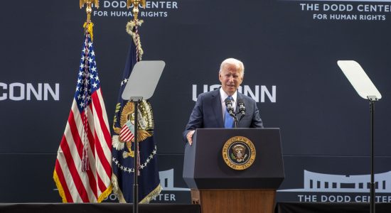 President Joe Biden speaking at The Dodd Center for Human Rights at the University of Connecticut. President Biden is standing at a podium featuring the U.S. Presidential seal with American flags in the background.