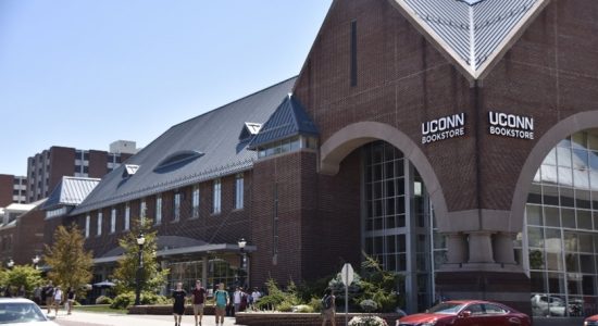 Exterior view of the UConn Bookstore building which is a large brick building at the corner of an intersection. Students walking on a sidewalk and cars driving through the intersection are in the foreground