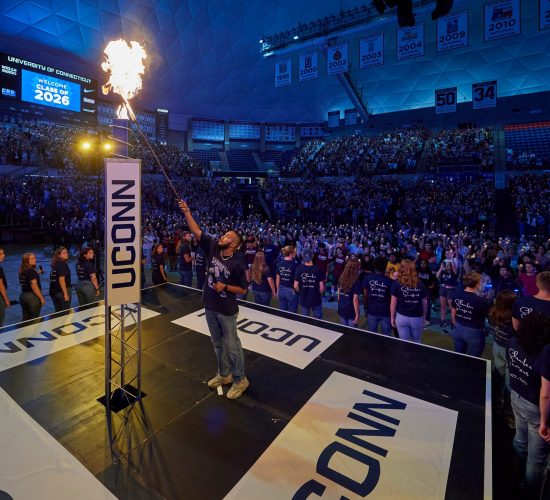 Individual lighting a large ceremonial torch on a stage surrounded by a crowd of students in Gampel Pavilion at the Class of 2026 Convocation event.