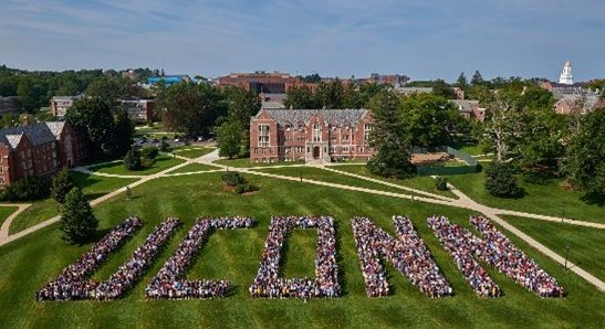 Aerial view of the Great Lawn at UConn with a large crowd standing so as to form the word "UCONN." Various brick buildings are visible in the background and sidewalks crisscross the perimeter of the lawn.