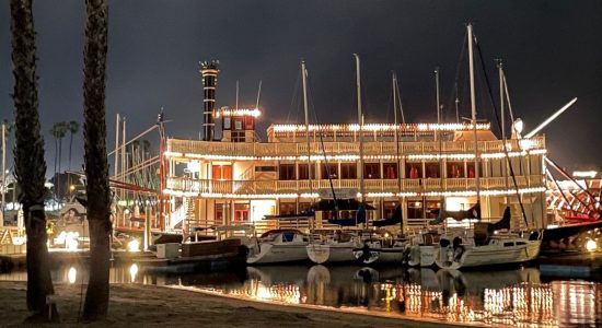 A riverboat in port lit up at night in San Diego, California. A variety of smaller sailboats are alongside the riverboat with a small beach in the foreground.