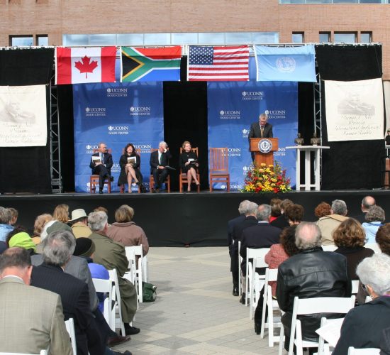 Dodd Prize dignitaries on an outdoor stage with various national flags hanging above the stage. A seated audience is in the foreground