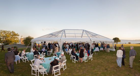 An outdoor banquet featuring a white canopy tent with open sides and table with light blue tablecloths and white chairs arranged outside the tent on grass. Guests are mingling in the tent and sitting at the outdoor tables.