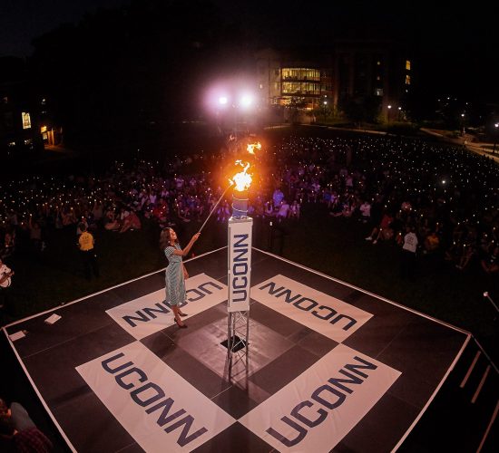 Individual lighting a large ceremonial torch on an outdoor stage during a Convocation ceremony