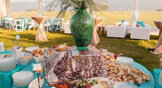 Cheese and vegetable platters on a table inside an outdoor tent. A seashore themed centerpiece is in the middle of the table which is covered in a teal blue tablecloth.