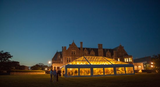 An outdoor tent at night illuminated from inside with guest milling about inside and outside the tent. In the background is the Branford Mansion on the UConn Avery Point campus