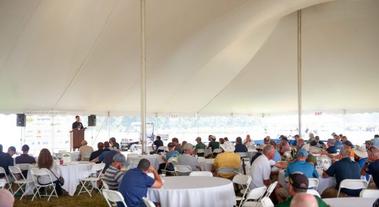 Outdoor banquet event during the 2022 UConn Turfgrass Conference. Guests are seated at tables under a large white tent listening to a speaker at a podium