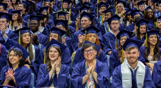 Graduates in UConn regalia seated in Gampel Pavilion applauding during a commencement ceremony