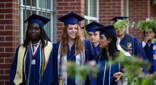 UConn graduates in blue and white caps and gowns processing outdoors to a commencement ceremony