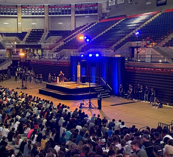 Students gathering around a stage in Gampel Pavilion listening to a speaker at a podium during Convocation