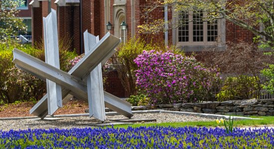 Abstract metal sculpture surrounded by blue flowers outside the Benton Museum of Art on the UConn Storrs campus. The red brick museum building is in the background