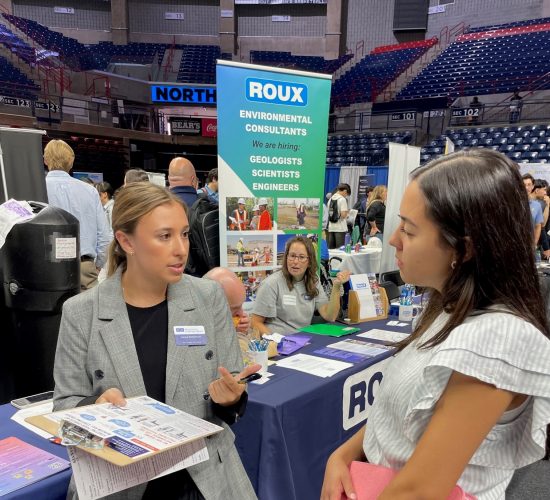 Professional representative speaking with a student visiting a booth at a career fair inside Gampel Pavilion