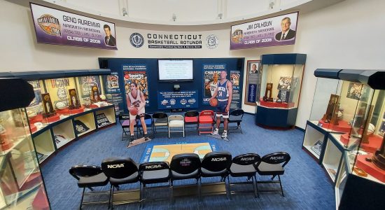 UConn Husky Sports museum multimedia interactive visitors area. Two rows of seats face a basketball display with a video screen and life-sized cut-outs of basketball players on either side of the screen. Along the walls are memorabilia displays.