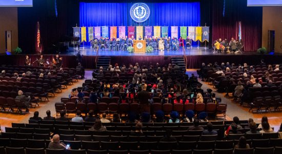 Awards ceremony on Jorgensen theater stage. Dignitaries and professors in regalia are seated on stage with various UConn school and college banners and an audience is seated in the foreground.