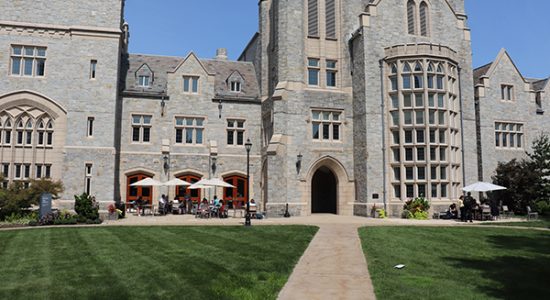 Exterior view of a gray stone gothic style building on the UConn Law campus. Patio tables with umbrellas are in front of the building and a sidewalk and green lawn is in the foreground