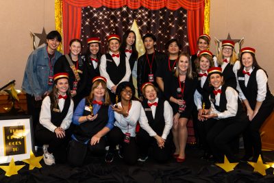 UECS staff and students dressed as ushers and paparazzi posing on the Movies and Musical themed stage at the 2023 Winter Holiday Celebration